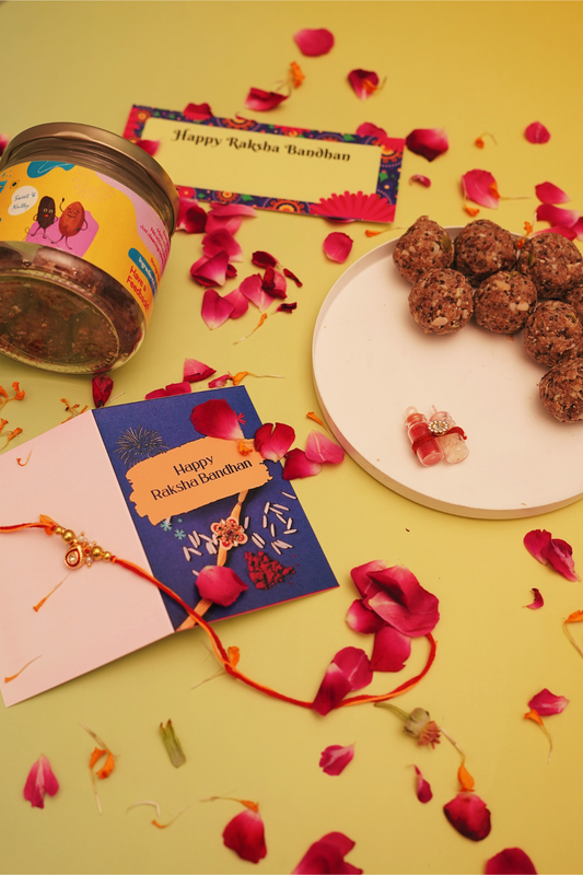 A styled flat-lay of a Raksha Bandhan gift set on a yellow background with scattered pink rose petals. The set includes a jar of seeds, a blue and pink greeting card, a white plate with brown rice balls, and a red thread bracelet.