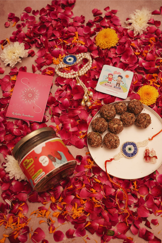 A styled flat-lay of a Raksha Bandhan gift set on a bed of red rose petals and marigold flowers. The set includes a jar of energy bites, a pink 'Raksha Bandhan' greeting card, an evil-eye rakhi and lumba, a fridge magnet, and a plate of energy bites with roli-chawal.