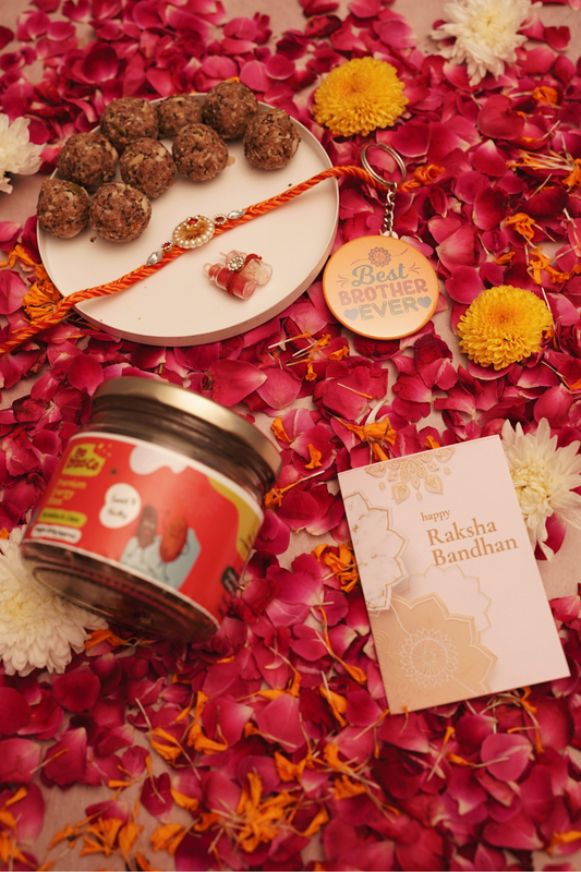 A styled flat-lay of a Raksha Bandhan gift set on a bed of red rose petals and marigold flowers. The set includes a plate of energy bites with a rakhi and roli-chawal, a 'Best Brother Ever' keychain, a jar of energy bites, and a peach 'Happy Raksha Bandhan' card.