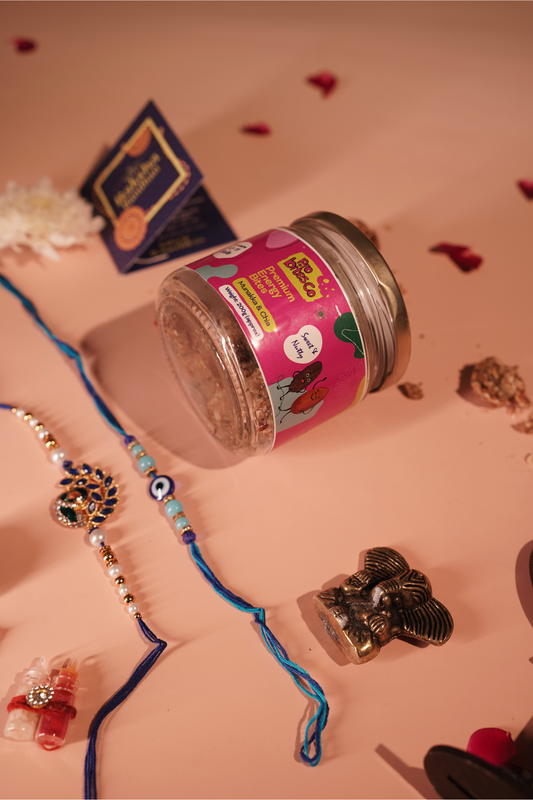 A jar of energy bites, a navy 'Raksha Bandhan' card, a peacock-design rakhi, an evil-eye rakhi, and a small brass Ganesh idol, styled on a peach background with scattered rose petals.
